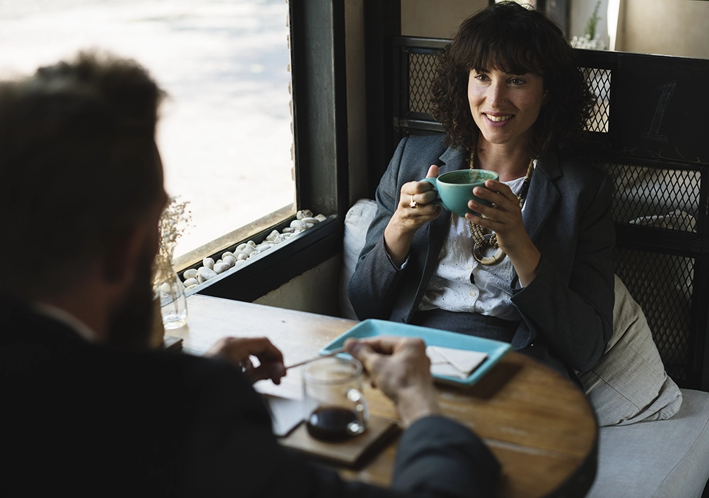 Woman in a blazer smiles while holding a teal mug at a round cafe table, talking with a man across from her.