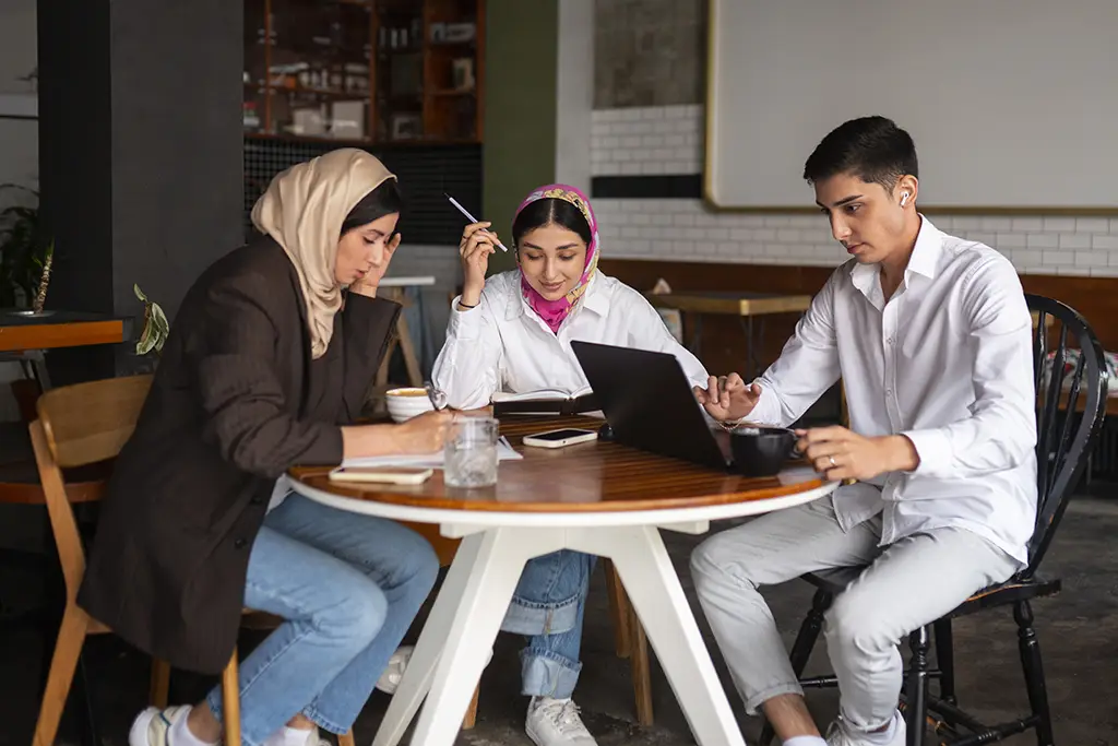 Three young adults study at a cafe table: two women in hijabs and a man with a laptop, papers and drinks on the table.
