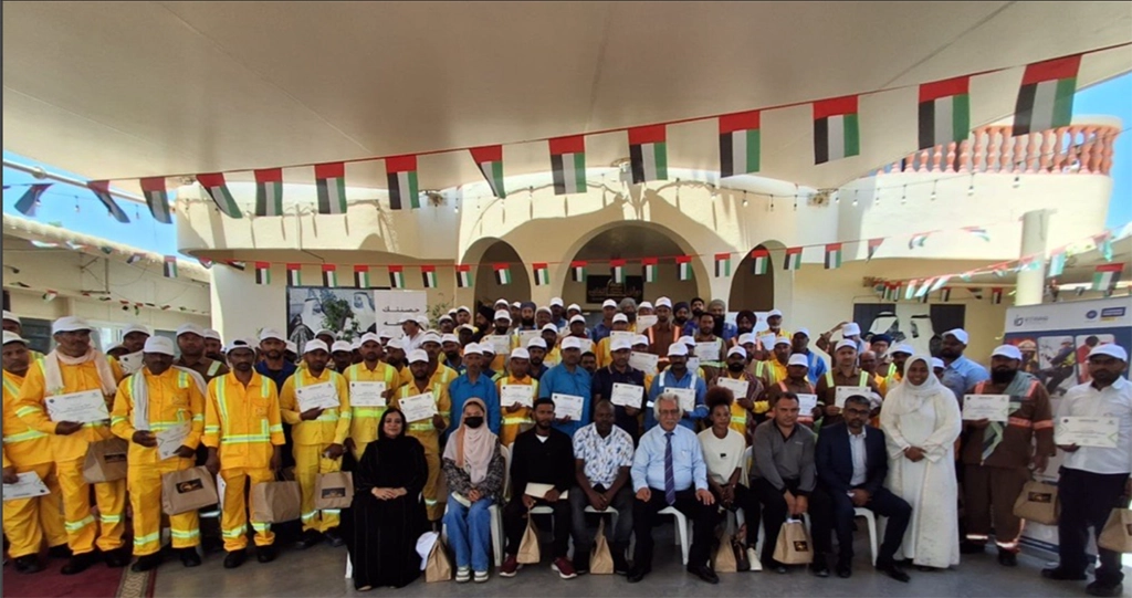 Group of workers in yellow safety uniforms posing with certificates at an outdoor ceremony under flag banners with a crowd behind them.