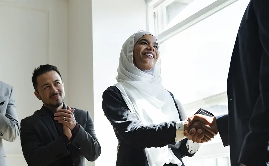 Professional woman wearing a white hijab shakes hands with a colleague as teammates applaud in a bright office setting.