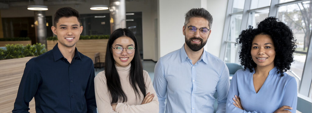 Four diverse colleagues stand together in a bright, modern office lobby, smiling at the camera.