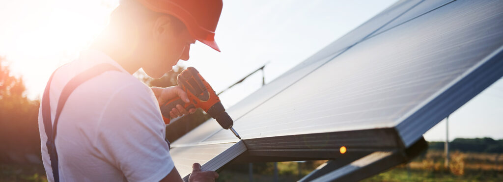 Worker in a red hard hat drills into a solar panel mounted on a tilted frame outdoors at sunset?