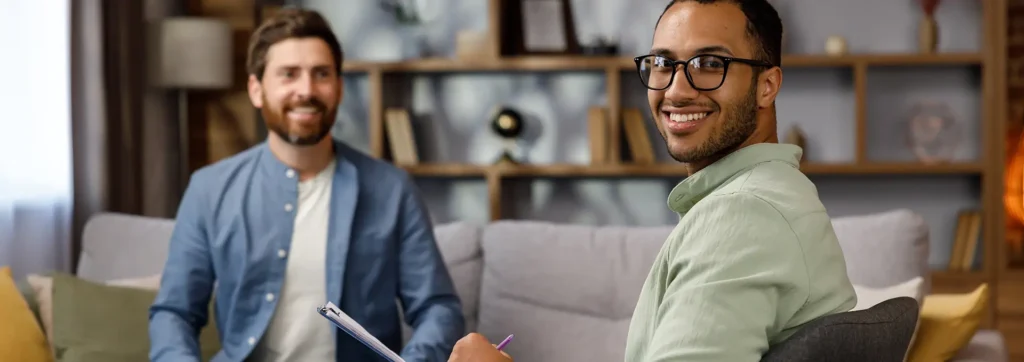 Two men sit on a couch in a cozy living room, smiling; one holds a clipboard and pen in a casual meeting.