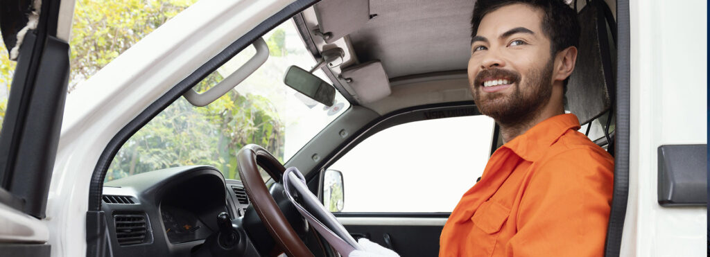 Smiling man in an orange work shirt sits in the driver's seat of a white truck cab.