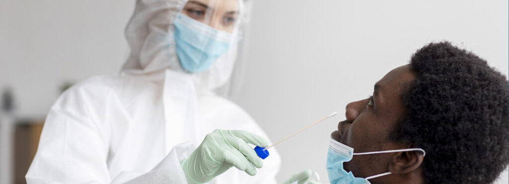 Medical professional in full PPE (gown, gloves, mask, face shield) collecting a nasal swab from a patient wearing a mask.