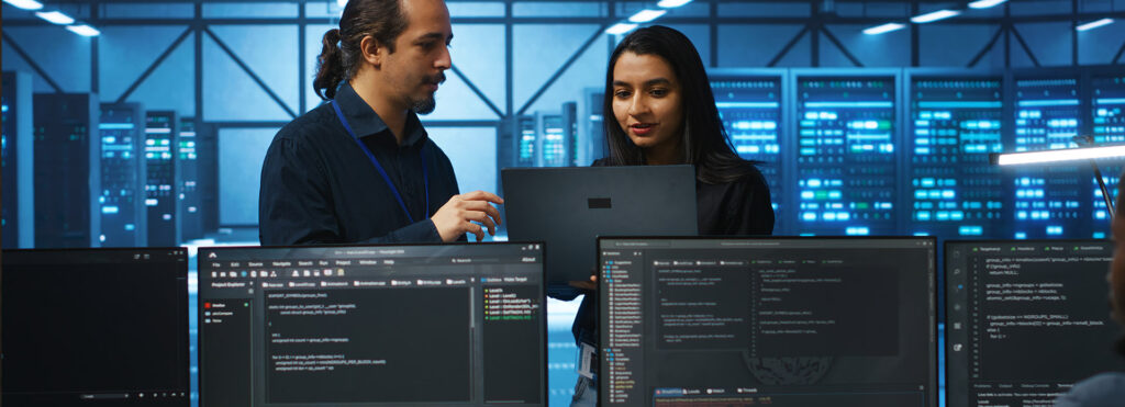 Two IT professionals review code on a laptop in a server room with blue-lit racks and multiple monitors showing code.