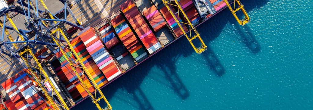 Top-down view of a cargo port with multicolored shipping containers and large yellow gantry cranes over turquoise water