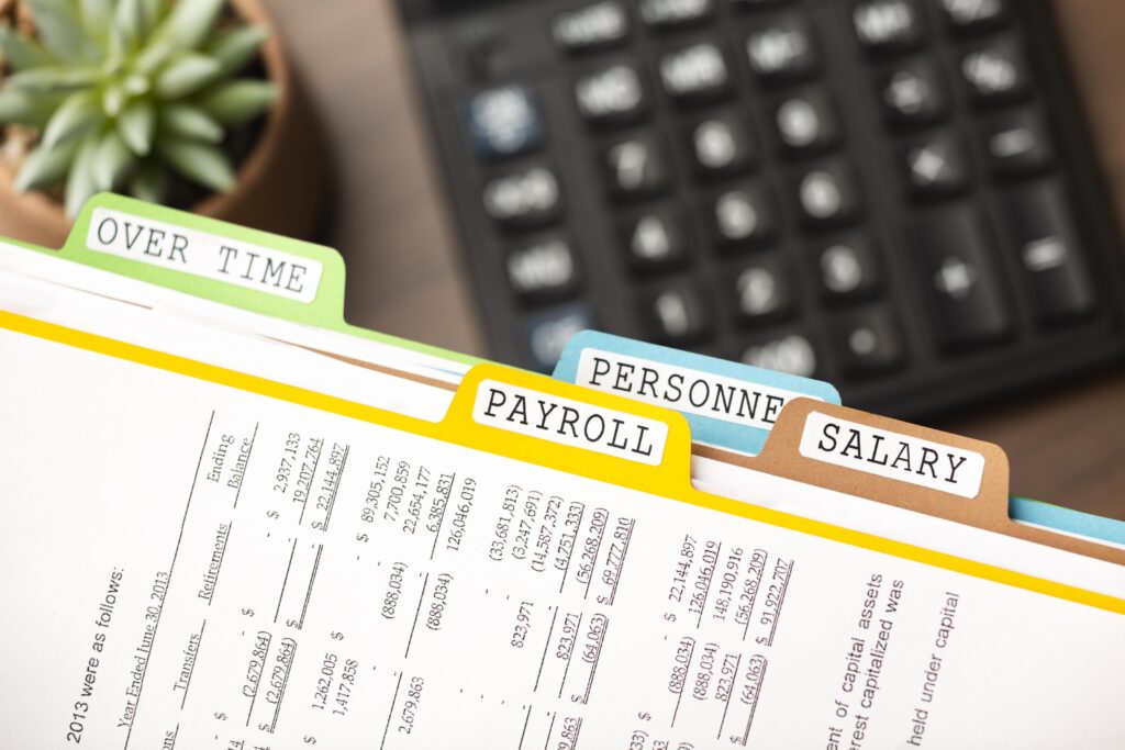 Colorful file folders labeled OVER TIME, PERSONNEL, PAYROLL, and SALARY sit on top of a printed financial document with a keyboard blurred in the background.