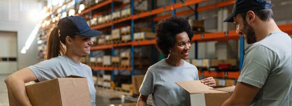 Three warehouse workers in gray shirts share a smile while handling cardboard boxes among tall orange shelving.